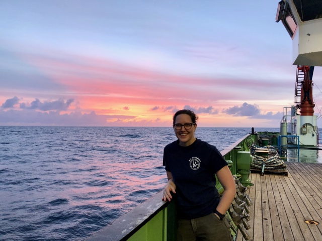 Erin Jones stands on the deck of a research vessel with the sea and sunset behind her