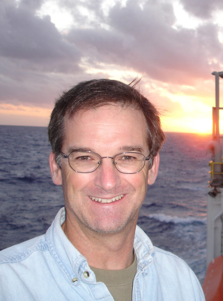 A head shot of one of the board members, Rick Murray, white males with dark short hair. The backdrop is the sun setting over the ocean. The corner of the stern of a research vessel can be seen in the right lower corner. 